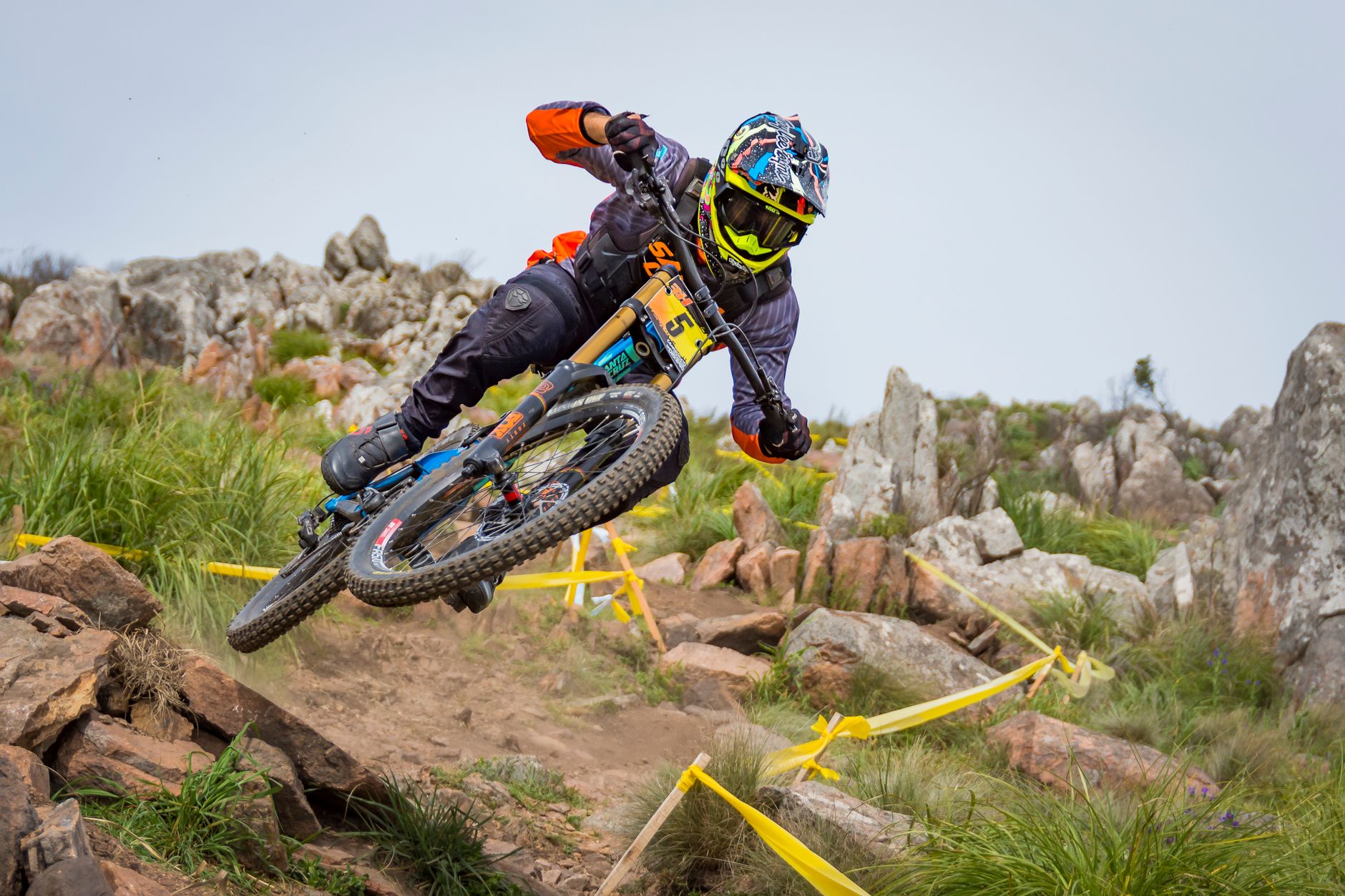 Downhill race bike with a dual-crown fork landing into a sharp rock garden on a taped race track.
