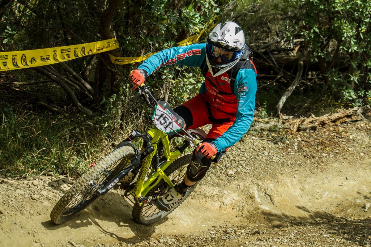 Downhill racer in a full-face helmet and race plate railing a dusty berm on a dual-crown race bike between course tape.