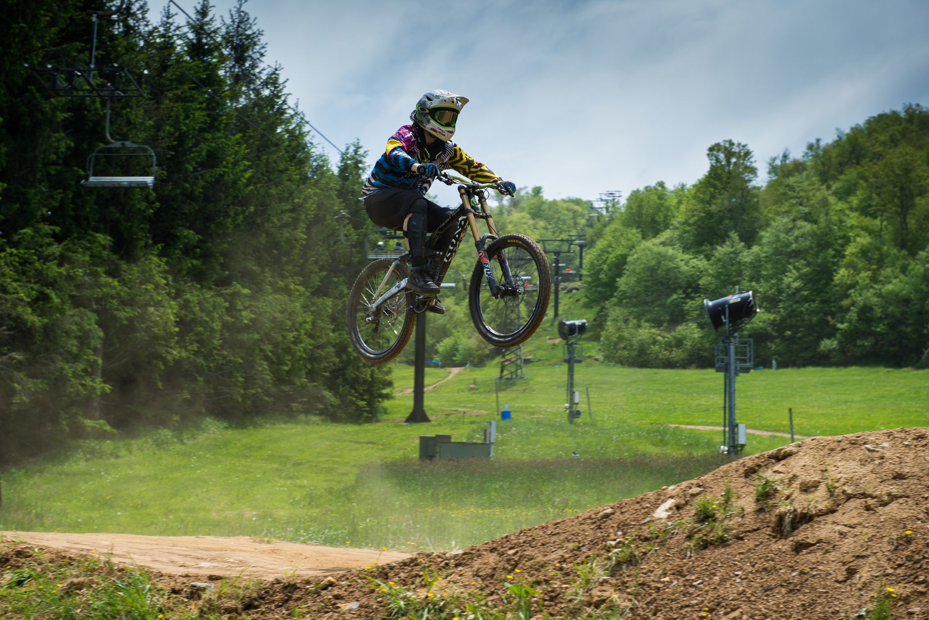 Downhill rider on a dual-crown Kona sending a dirt jump on a lift-served bike park trail, chairlift towers behind.