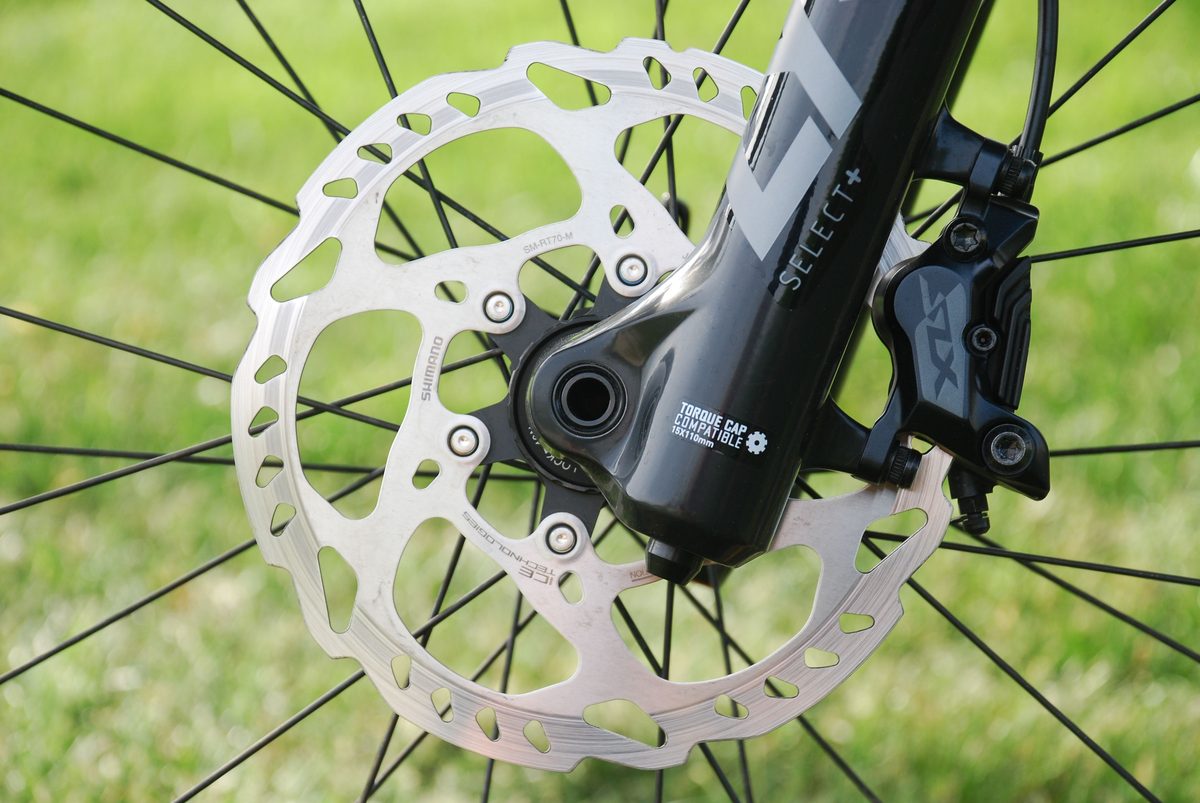 Close-up of a RockShox fork lower leg labelled Select+ and Torque Cap Compatible, beside a Shimano SLX brake caliper and rotor.
