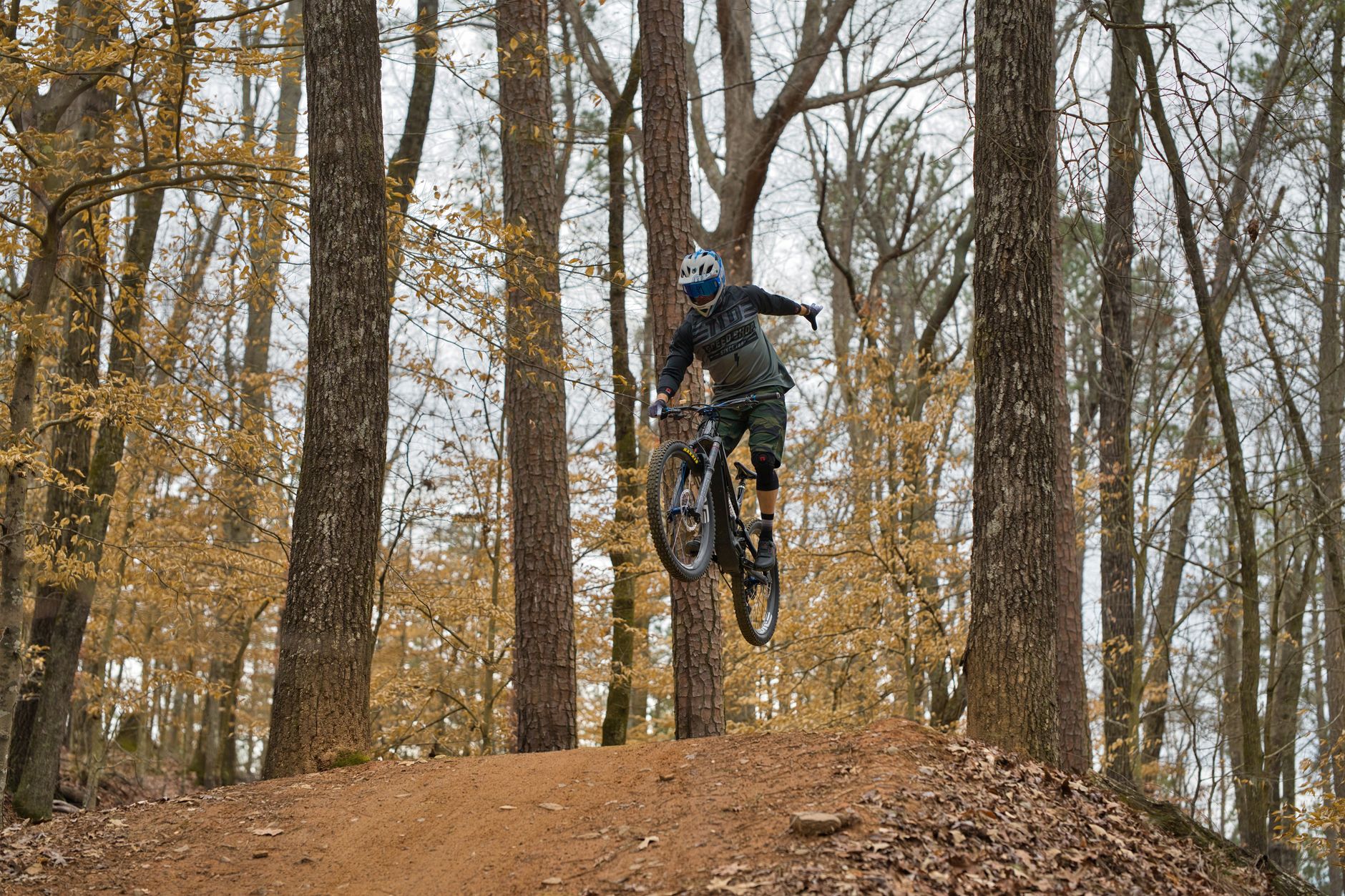 Rider in a full-face helmet jumping a full-suspension mountain bike on a dirt lip in an autumn forest.