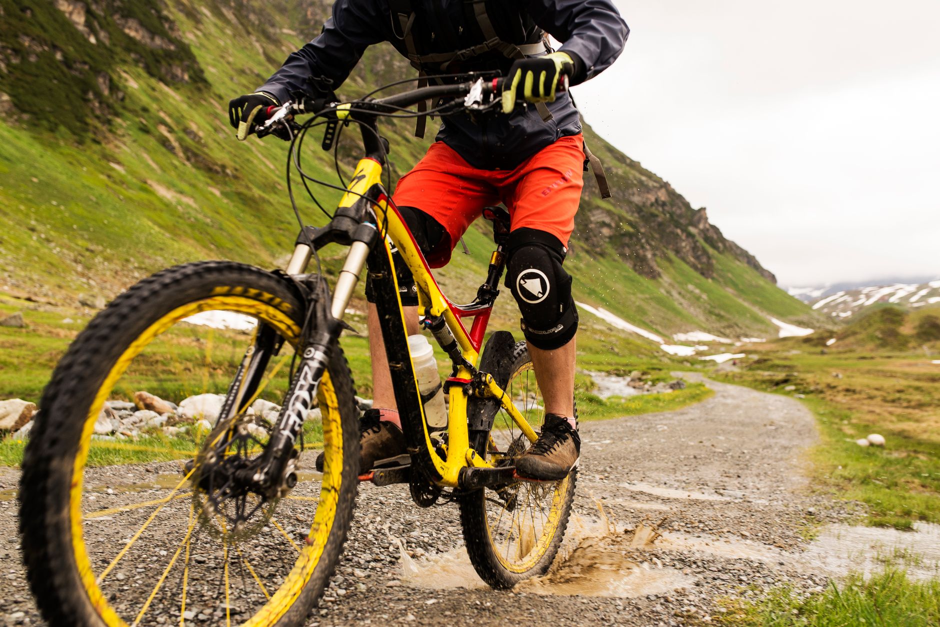 Rider on a yellow full-suspension trail bike splashing through standing water on a wet alpine trail.