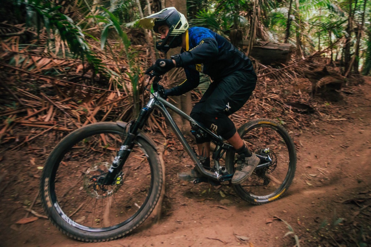 Full-suspension enduro mountain bike carving a loamy forest corner, rider in full-face helmet.