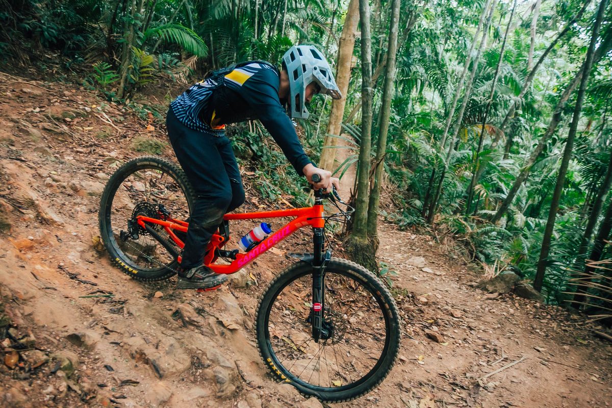 Rider descending a steep, rocky jungle trail on an orange Santa Cruz full-suspension bike with Maxxis tires.