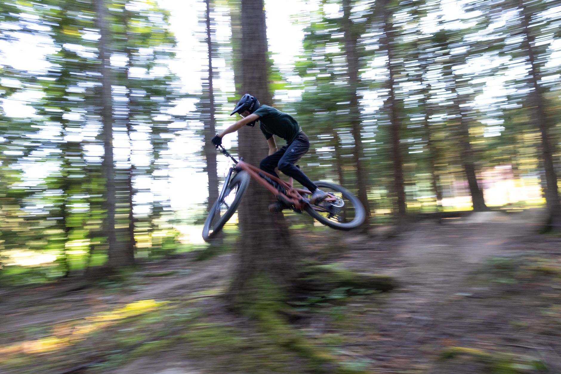 Rider mid-jump on a full-suspension trail bike, body low and centred over the frame on a forest trail.