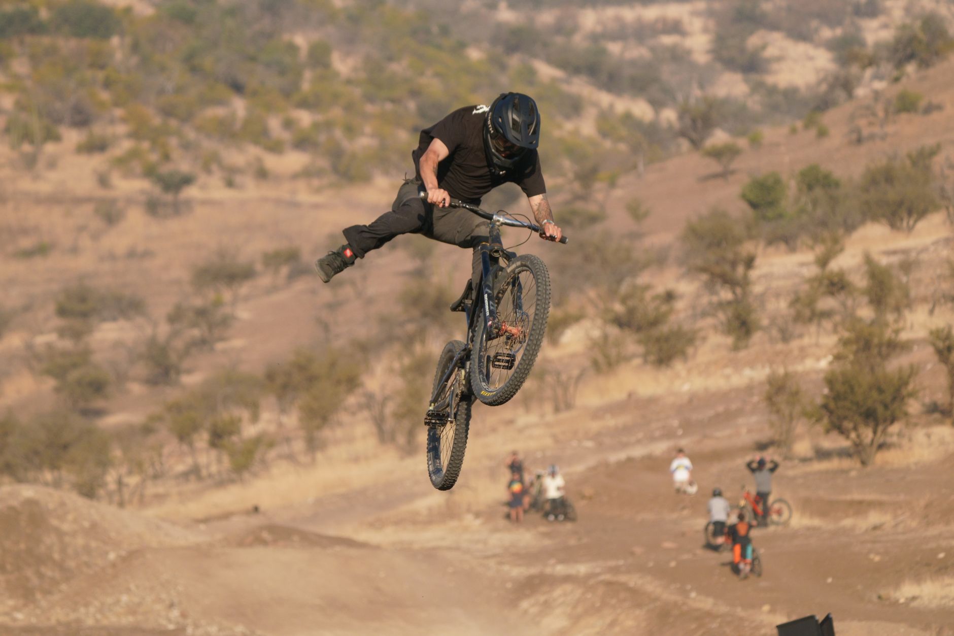 Rider throwing a no-hander on a full-suspension bike above a dirt jump in dry, hilly desert terrain.
