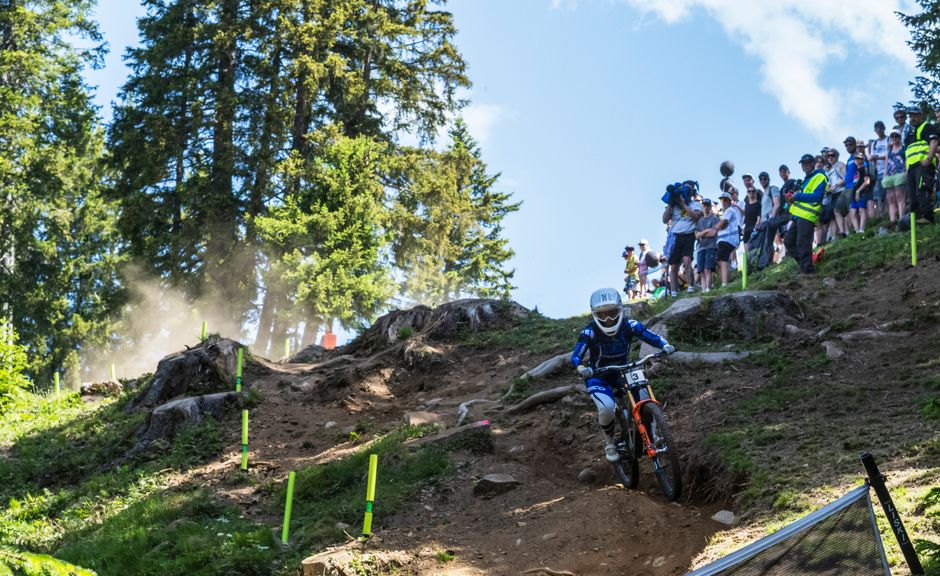 Downhill racer in a full-face helmet dropping into a steep, dusty race track between tape and spectators in Switzerland.