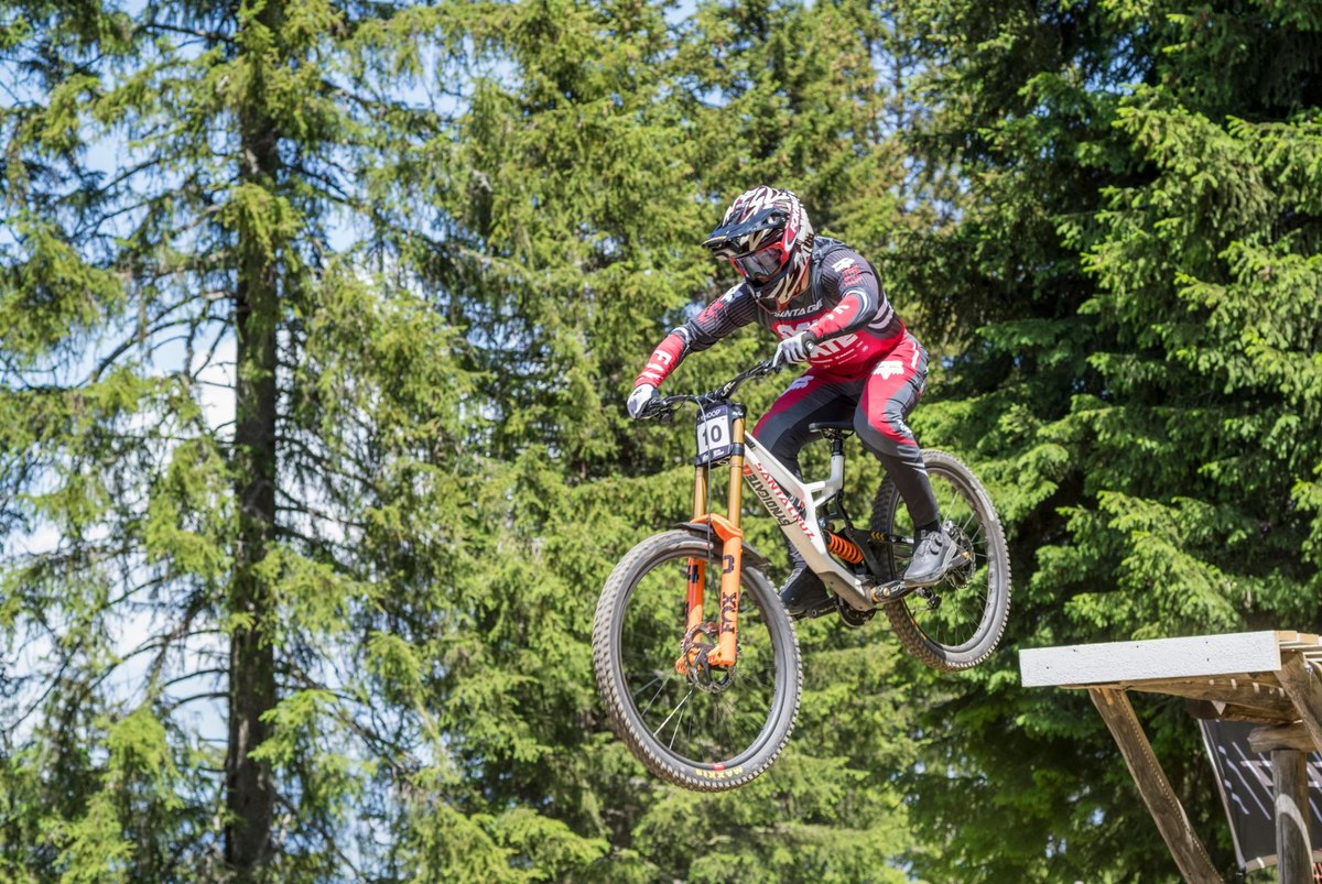 Downhill racer with a race plate mid-air off a wooden jump on a Santa Cruz V10, orange dual-crown Fox 40 fork in full view.