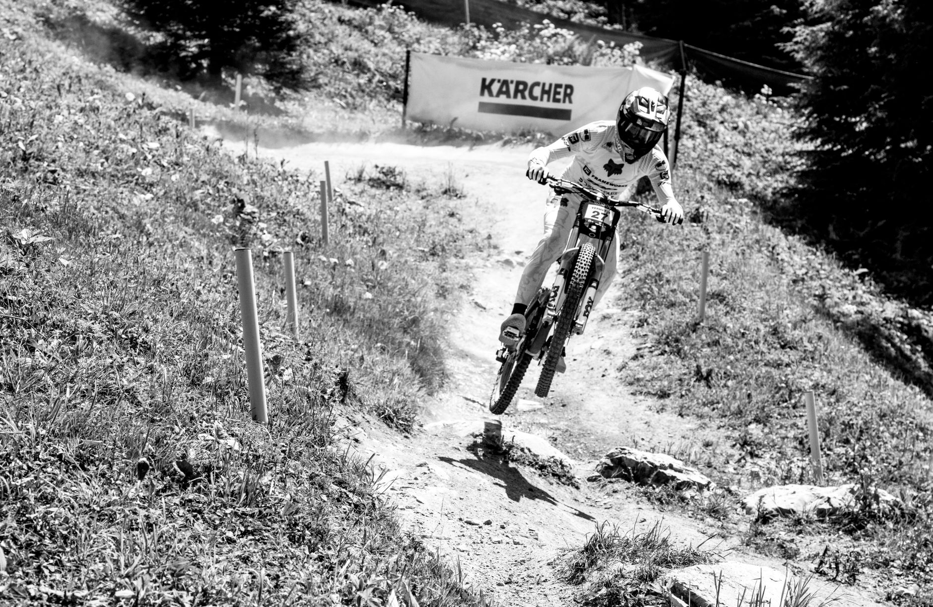 Black and white photo of a rider airborne over a small jump on a downhill bike on a dry, rocky trail.