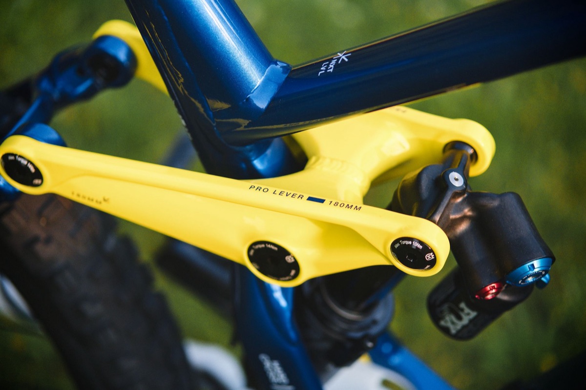 Close-up of a yellow rocker link driving a Fox rear shock on a blue full-suspension mountain bike frame.