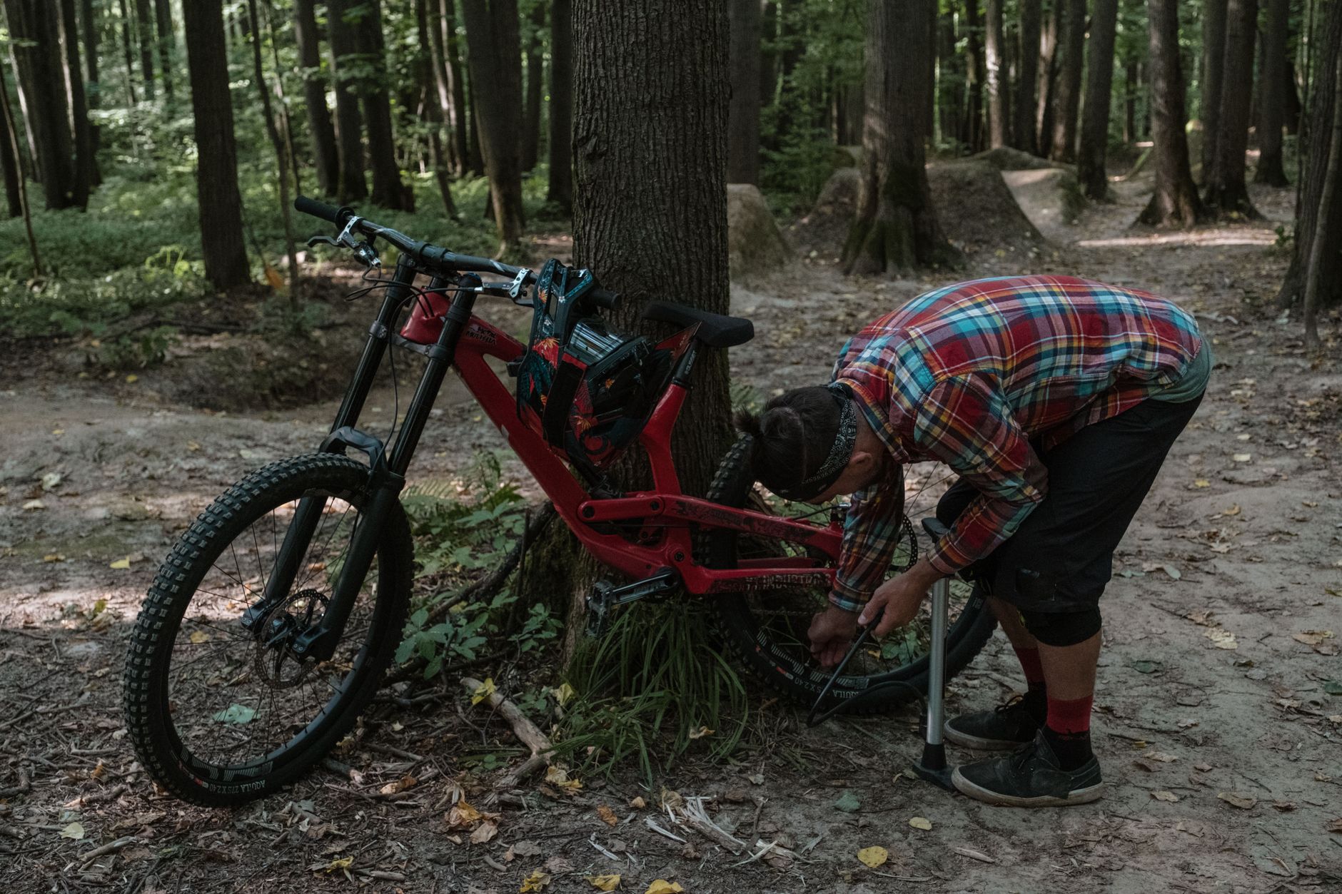 Rider in a plaid shirt bent over a red downhill mountain bike leaning against a tree beside a dirt jump trail in the woods.