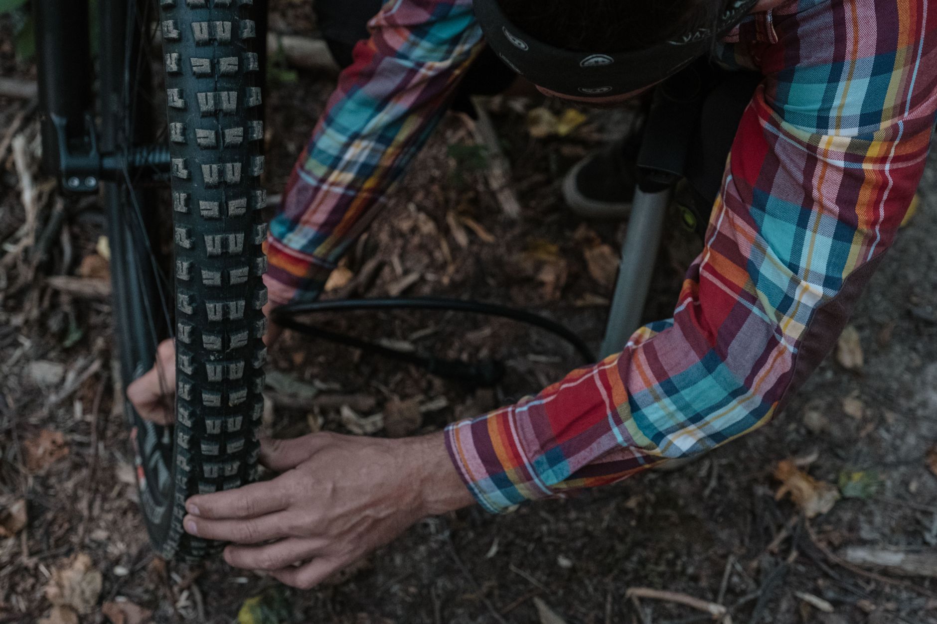 Hands squeezing the knobby front tire of a mountain bike on a forest trail, with a floor pump hose beside it.
