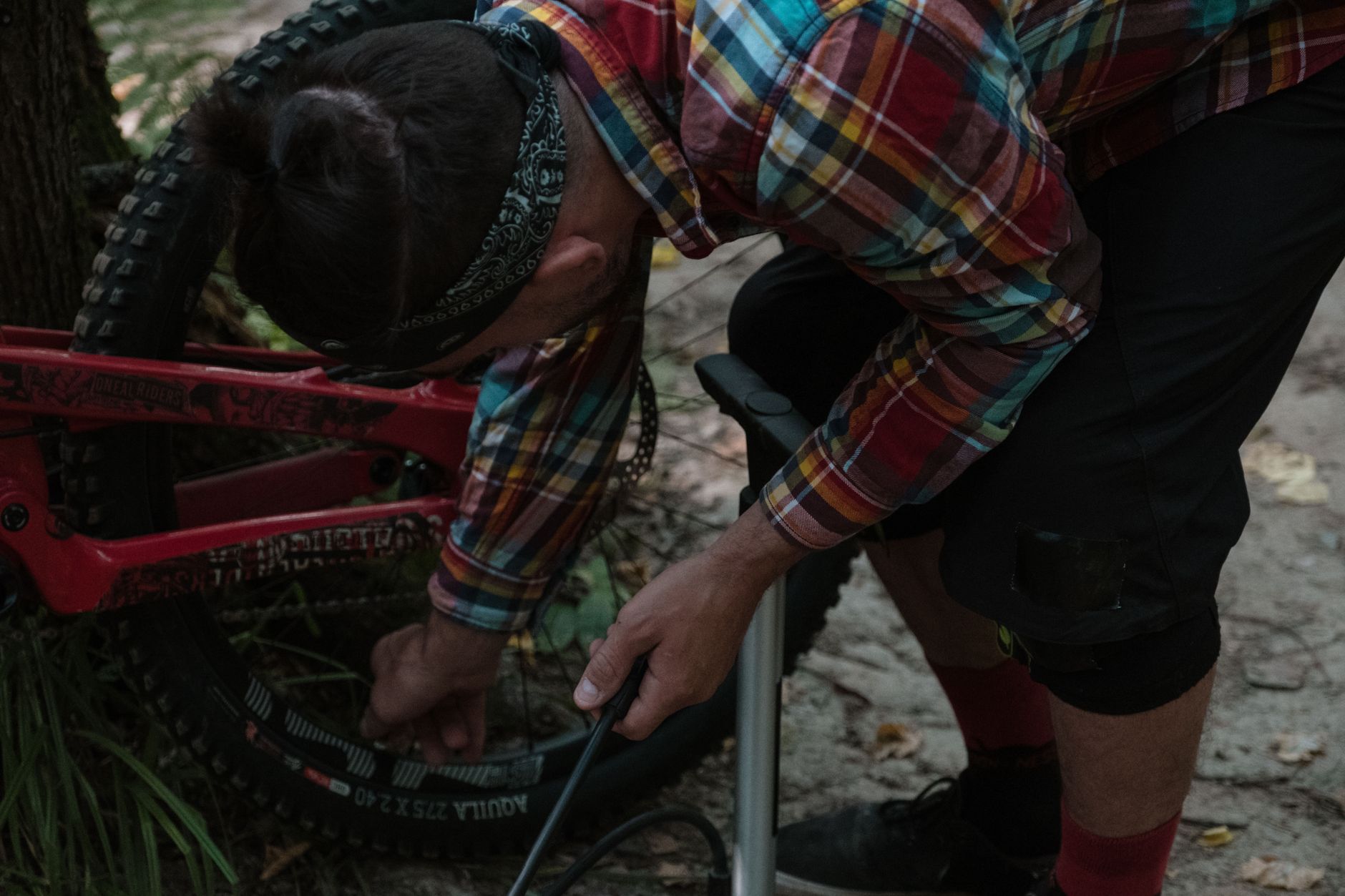 Rider crouched beside a red full-suspension mountain bike on a dirt trail, pumping up a Maxxis 27.5 x 2.40 tire with a floor pump.
