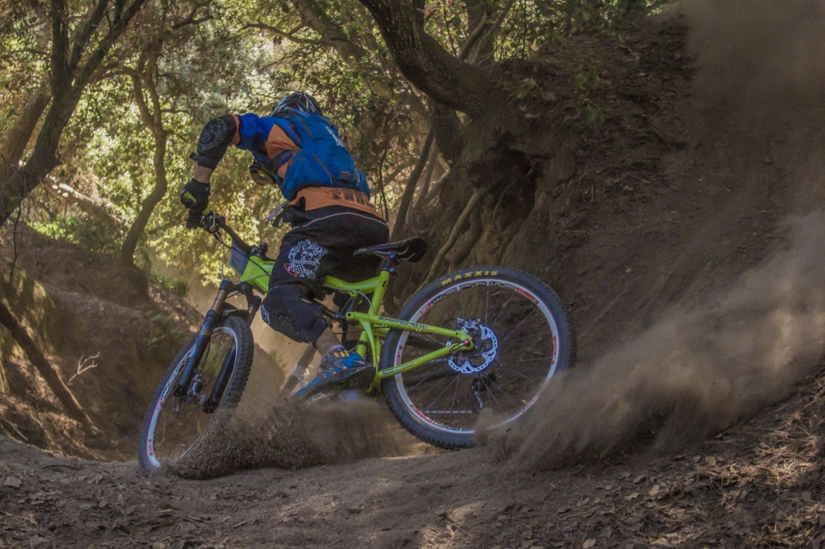 Rider drifting a full-suspension mountain bike through a dusty forest corner, rear wheel breaking loose.