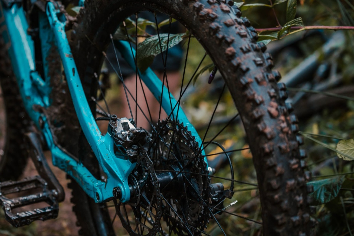 Rear end of a muddy full-suspension trail bike, Shimano XT cassette, caliper, and hub in focus.